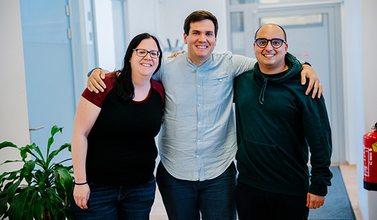 Ein Mann in der Mitte mit kurzem dunklem Haar und blauem Hemd, umarmt links eine Frau mit schwarzem T-Shirt, Brille und langem, braunem Haar, und rechts einen Mann mit kurzem, dunkelbraunem Haar, Brille und dunkelgrünem Pullover