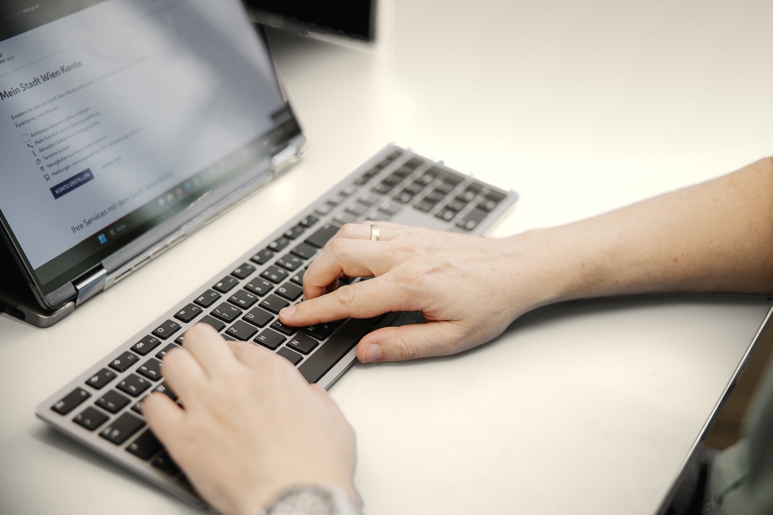 Hände tippen auf einer silbernen Tastatur an einem hellen, cremefarbenen Schreibtisch. Daneben steht ein aufgeklappter Laptop, auf dem die Seite „Mein Stadt Wien Konto“ geöffnet ist.